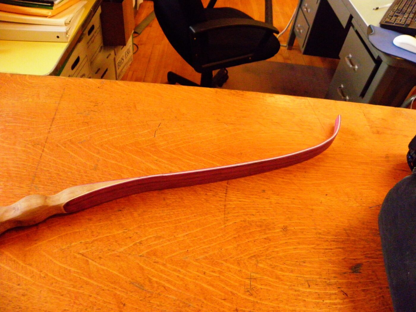 A wooden and red recurve bow lies on a workbench indoors.