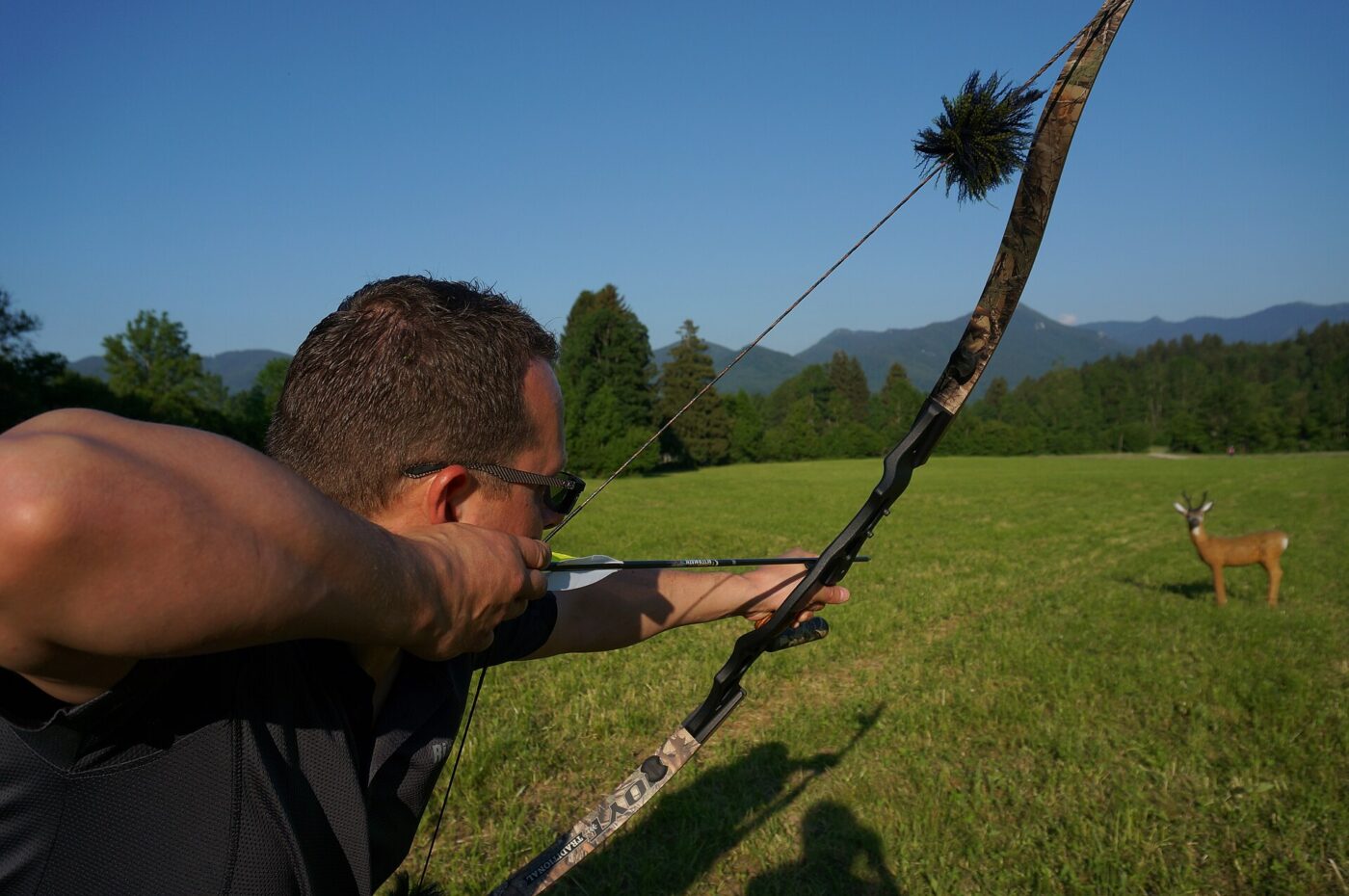 A man in an open field draws a recurve bow to full extension, aiming at an unseen target.