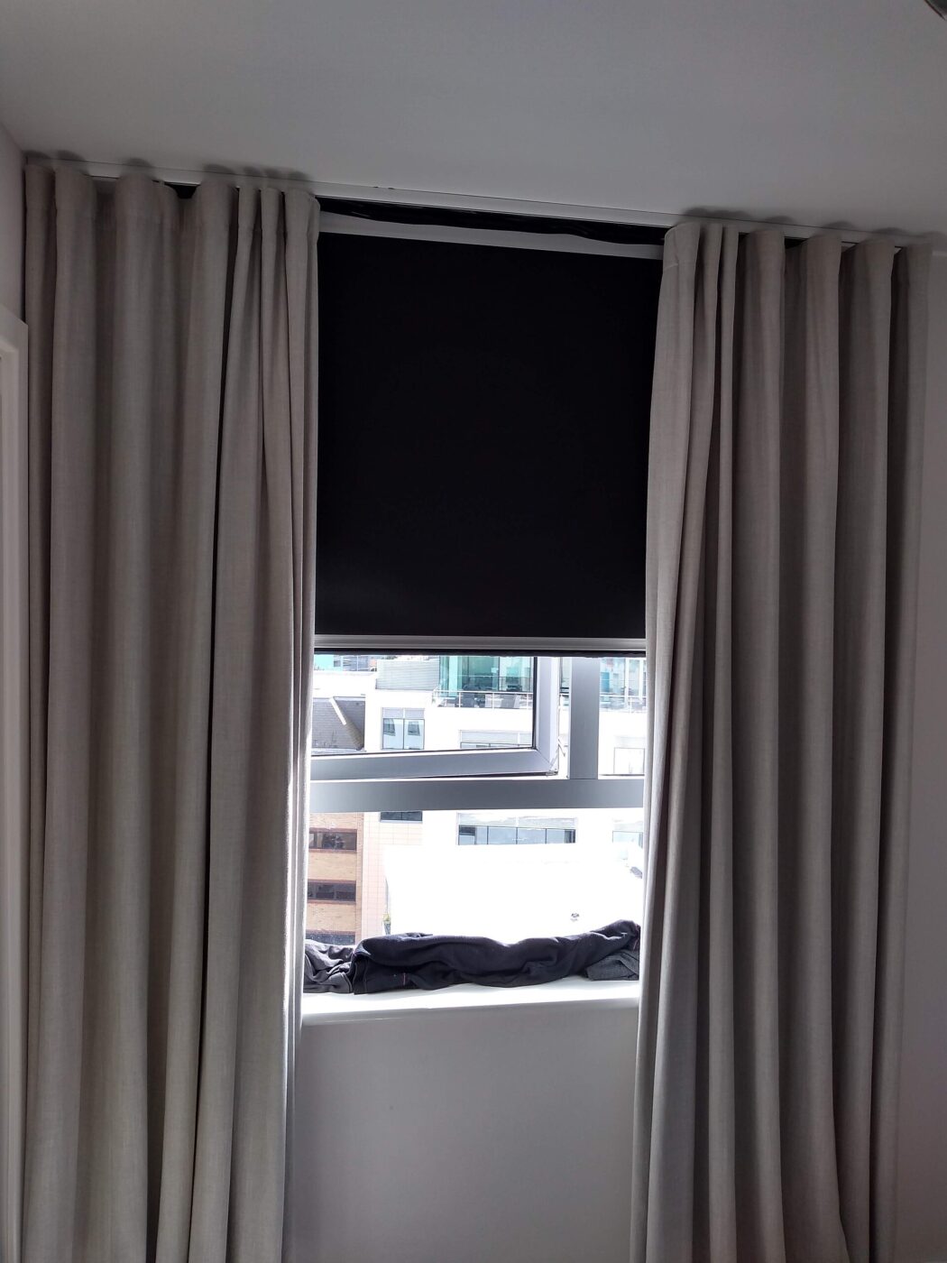 A dark roller blind closed over a window, framed by curtains.