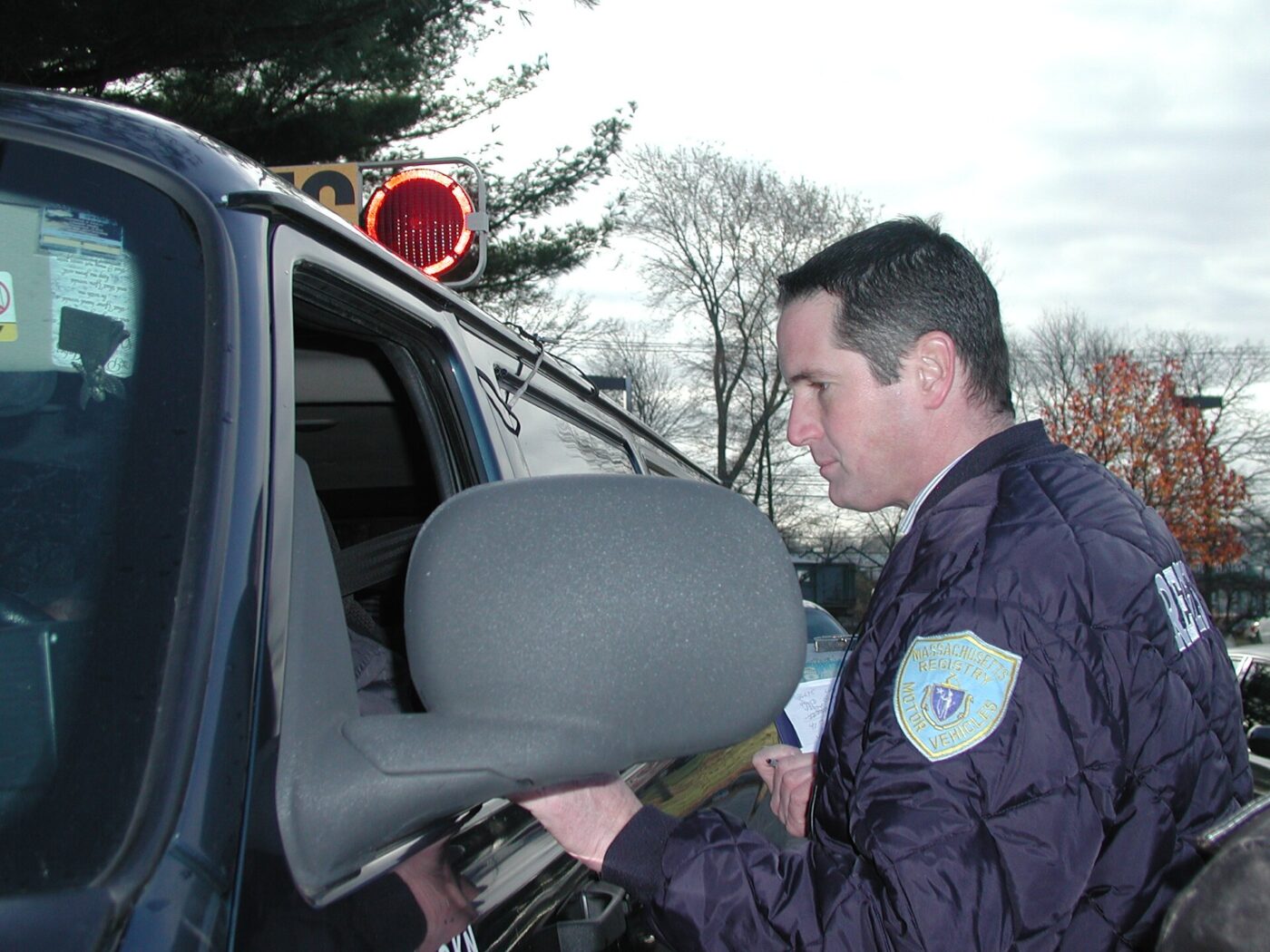 An inspector examines a rear-facing infant car seat beside an open vehicle door.