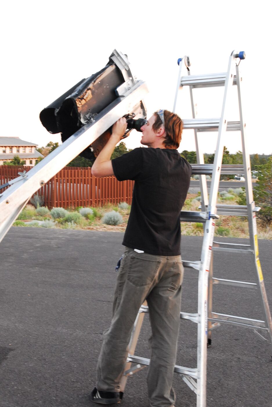 A person standing on a step ladder to look through a tall telescope outdoors.
