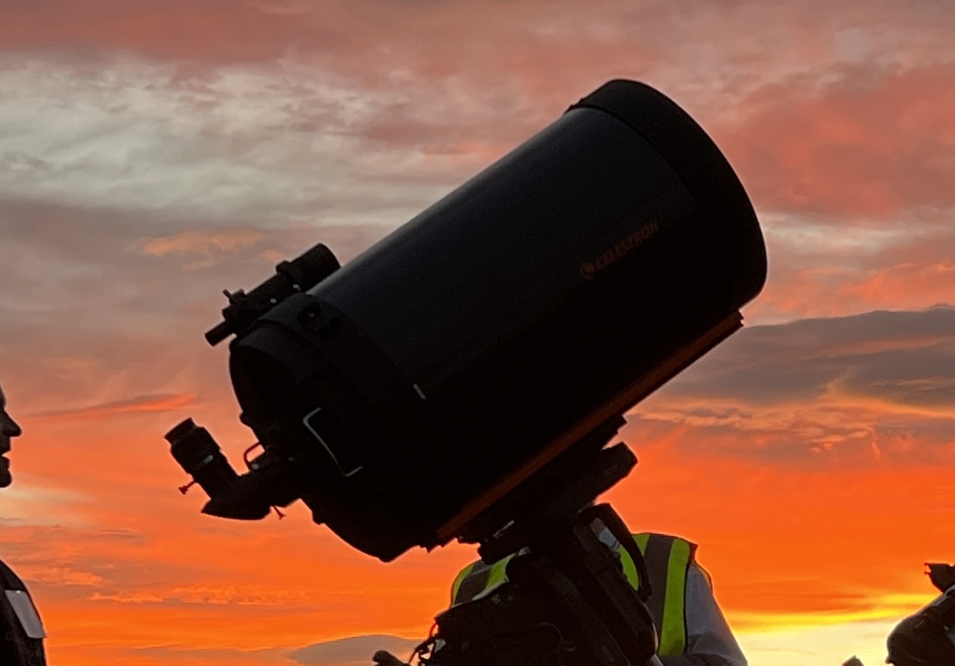 A silhouetted telescope on a tripod set up outdoors against an orange sunset.