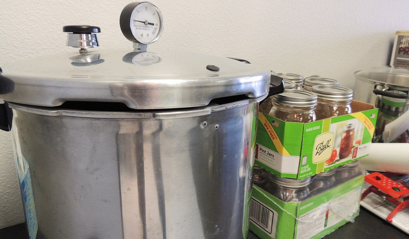 Stainless pressure canner with a dial gauge lid beside stacked canning jars.