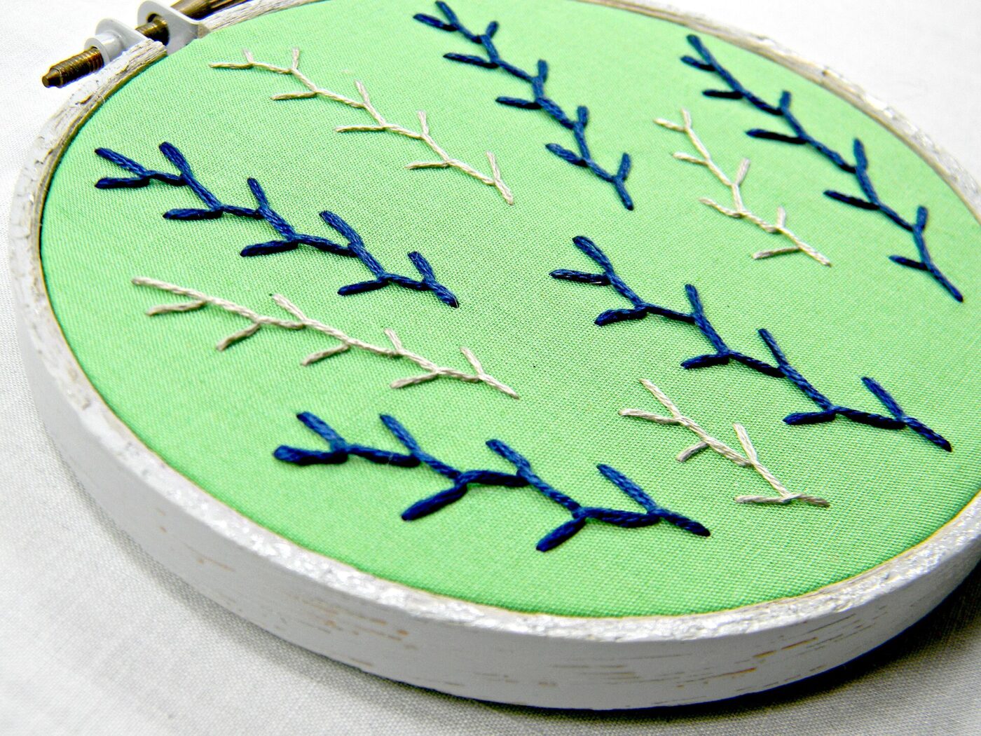 A round wooden embroidery hoop holding green fabric stitched with a blue feather pattern.