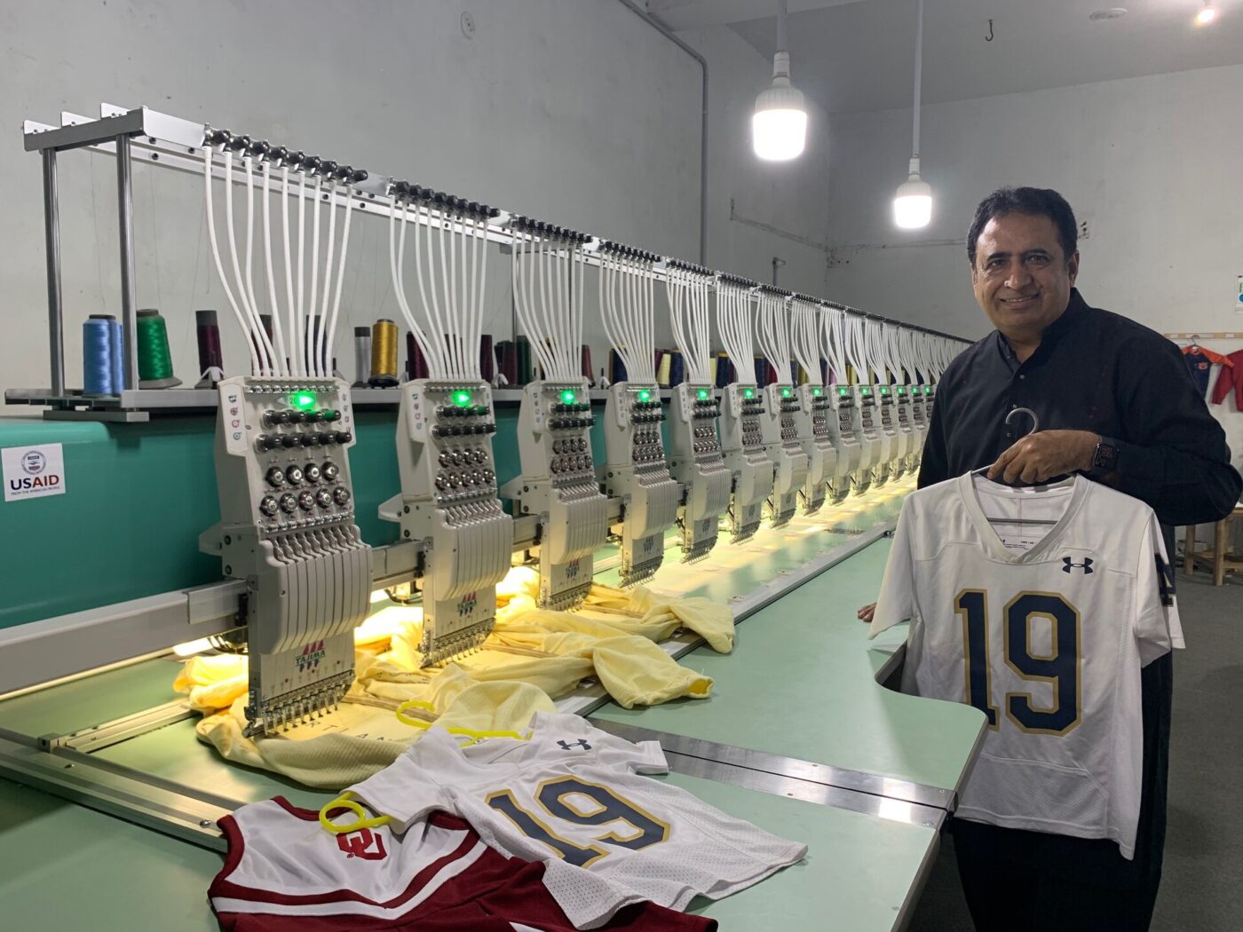 A man stands next to a long industrial embroidery machine with many thread spools stitching a shirt.