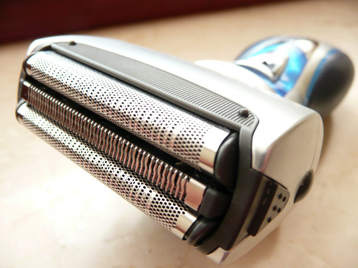 Close angled view of a silver and blue cordless electric shaver with two perforated metal foil heads and a center trimmer strip.