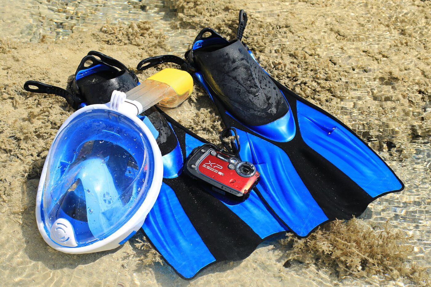 A blue full-face snorkel mask with an integrated tube on wet sand beside black-and-blue fins and a red camera.
