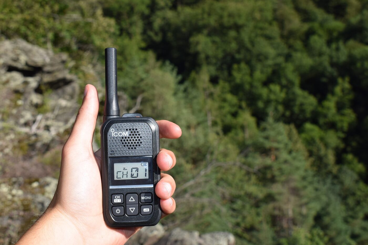 A person's hand holds a black handheld two-way radio while standing on a rocky cliff outdoors.