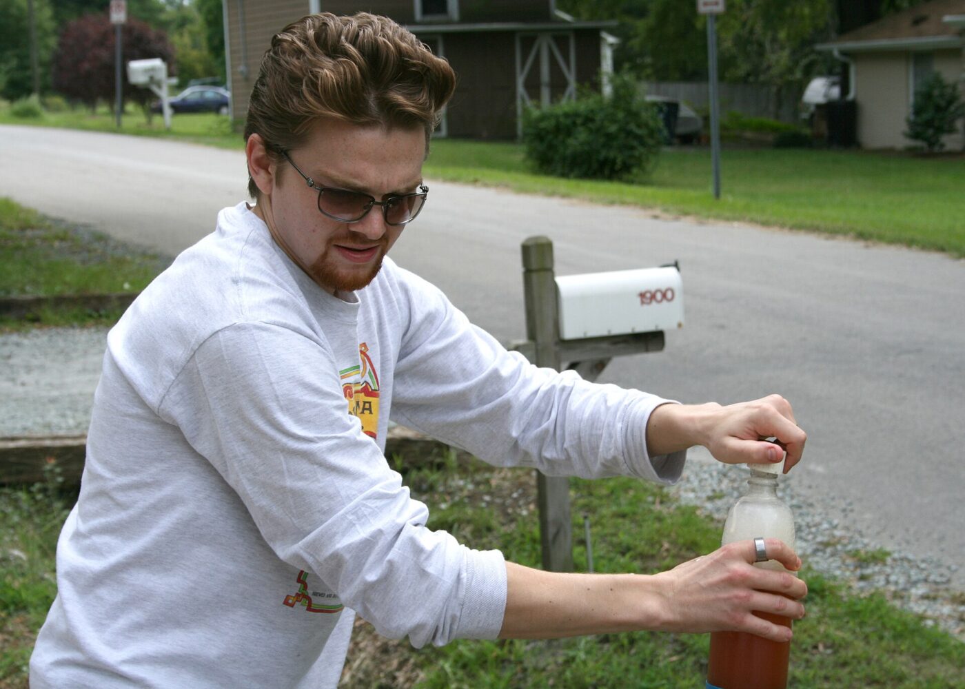 A man in sunglasses reacts while opening a capped bottle of homebrewed beer outside near a mailbox.