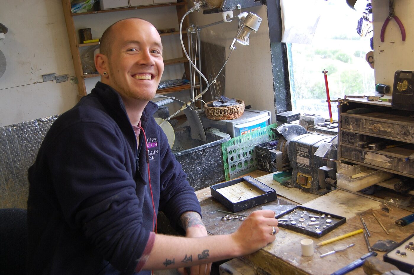 A jeweller at his cluttered workbench. That bench is what a handmade premium pays for.