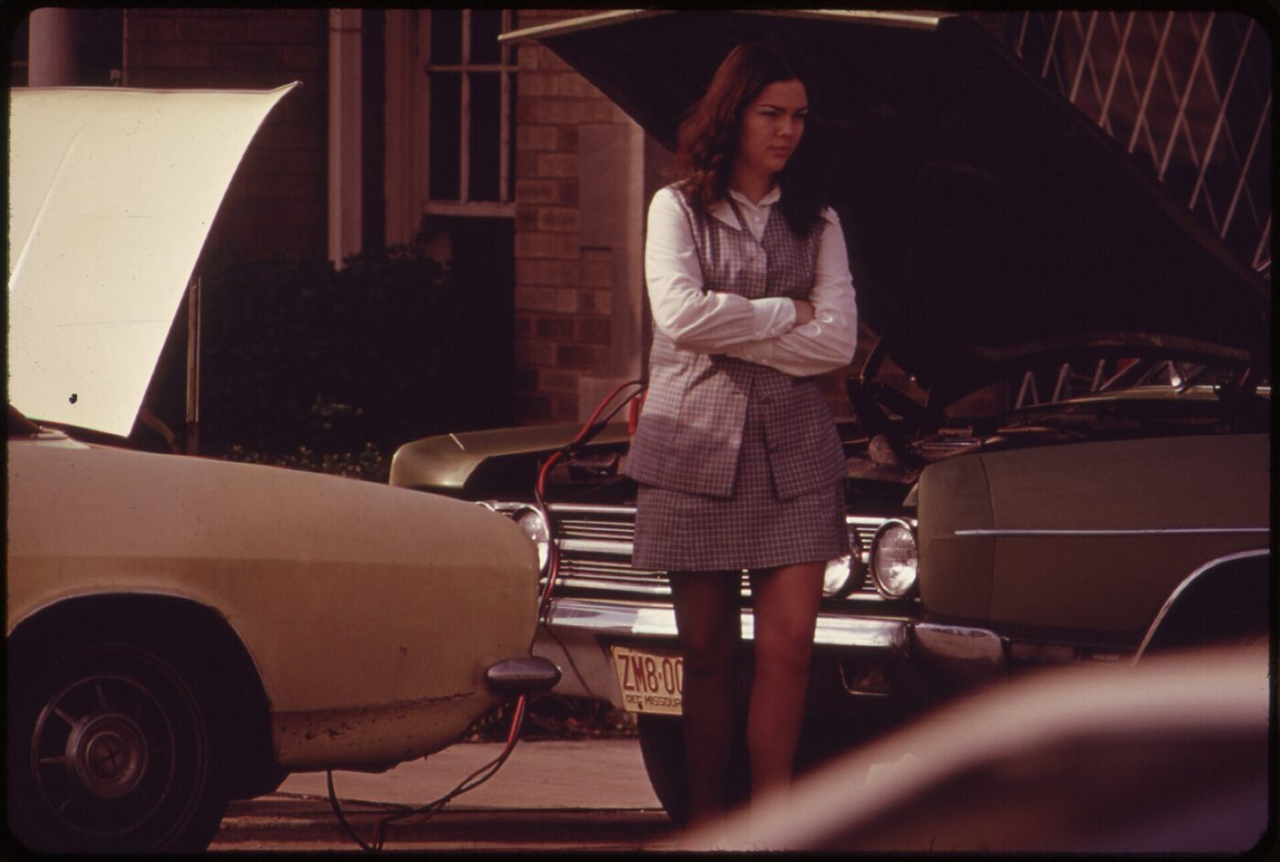 A commuter waits beside her car during a jump-start from another car, Chicago, 1970s.