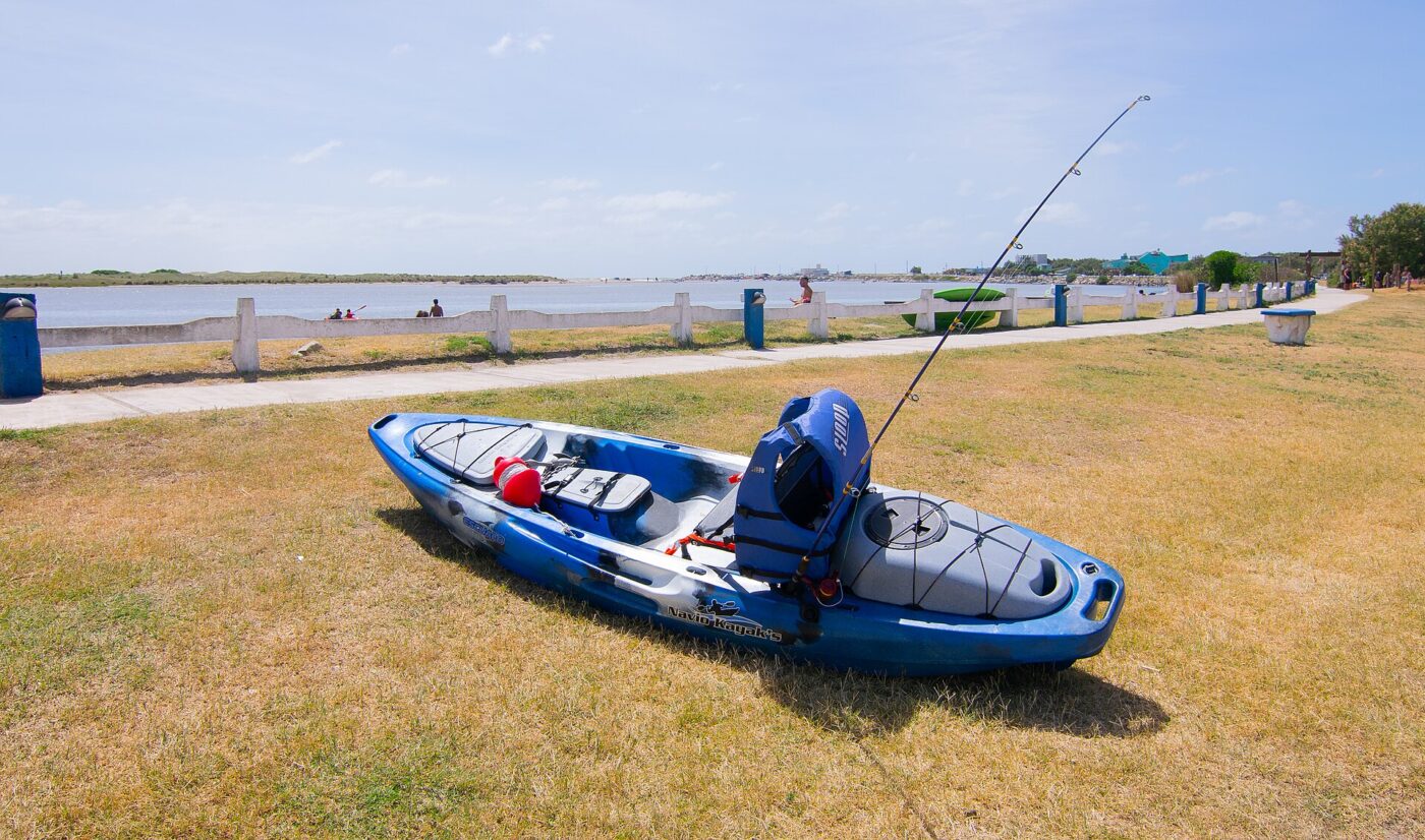 A blue and white fishing kayak with rod holders sits on grass near a paved boat launch.