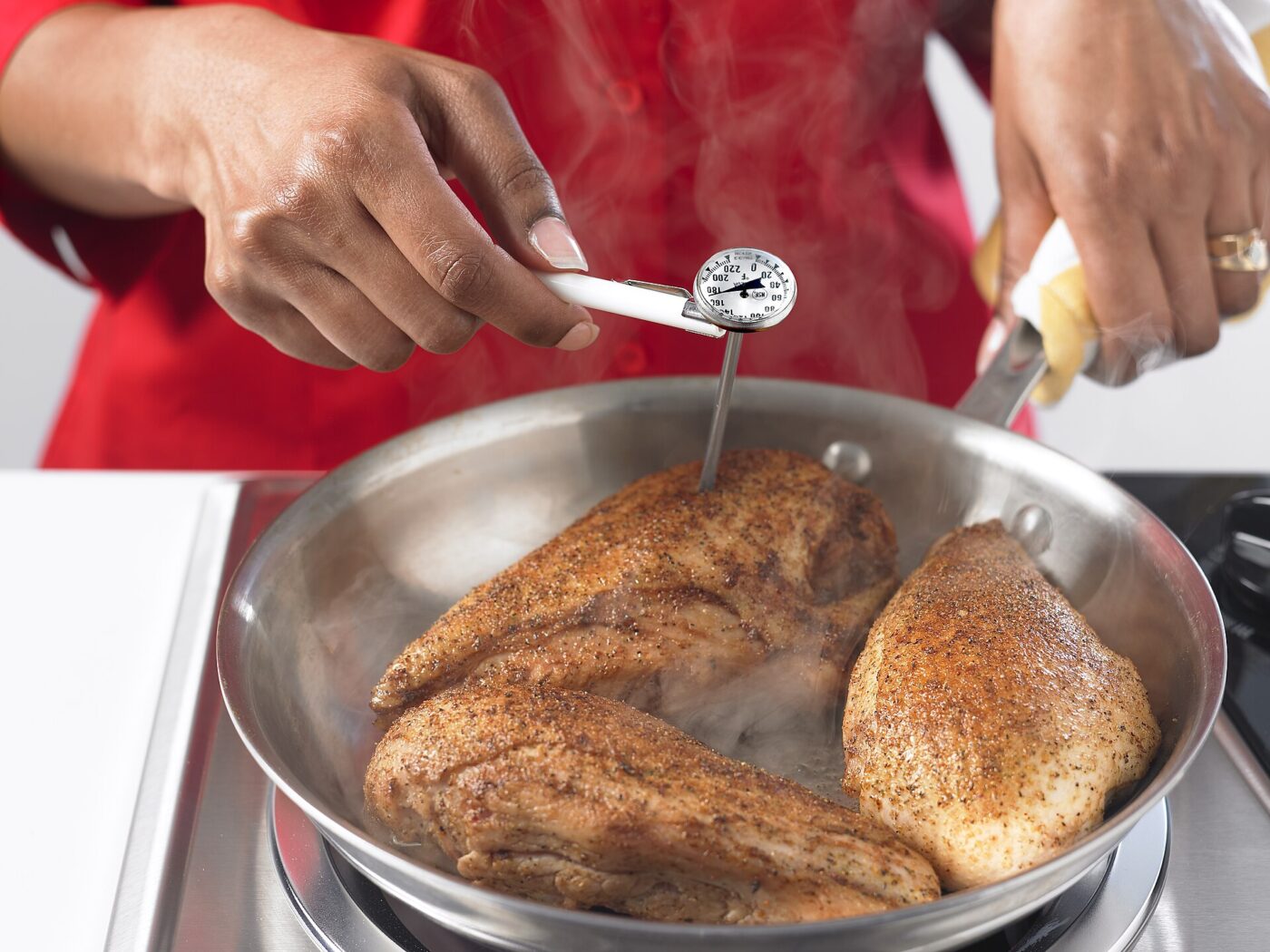 A person in a red apron inserts a dial probe thermometer into a piece of meat in a pot.