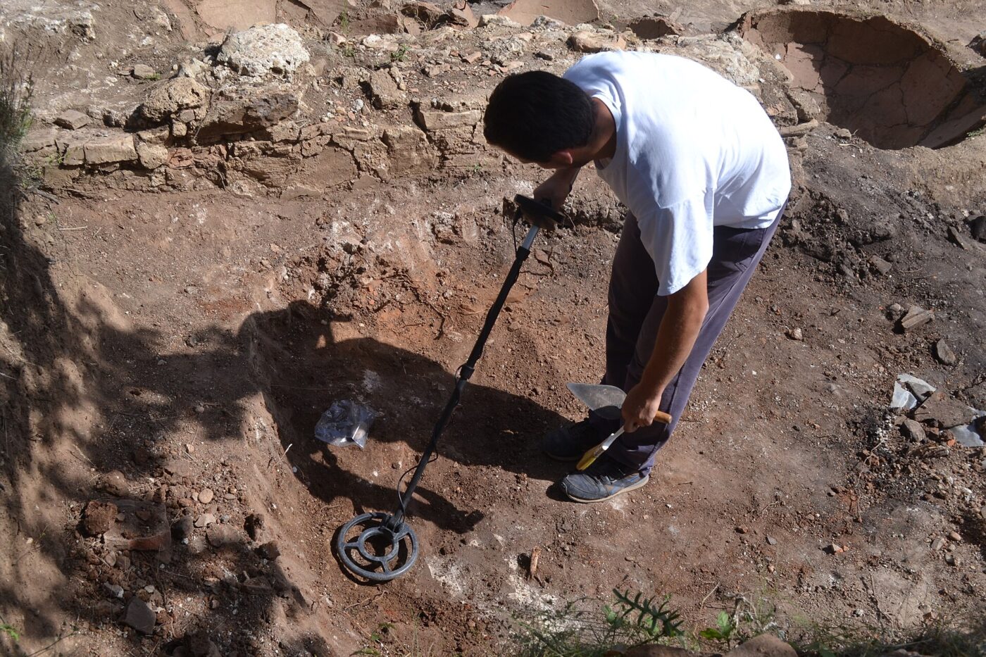 A person crouched over a hole in bare dirt, digging with a metal detector nearby.