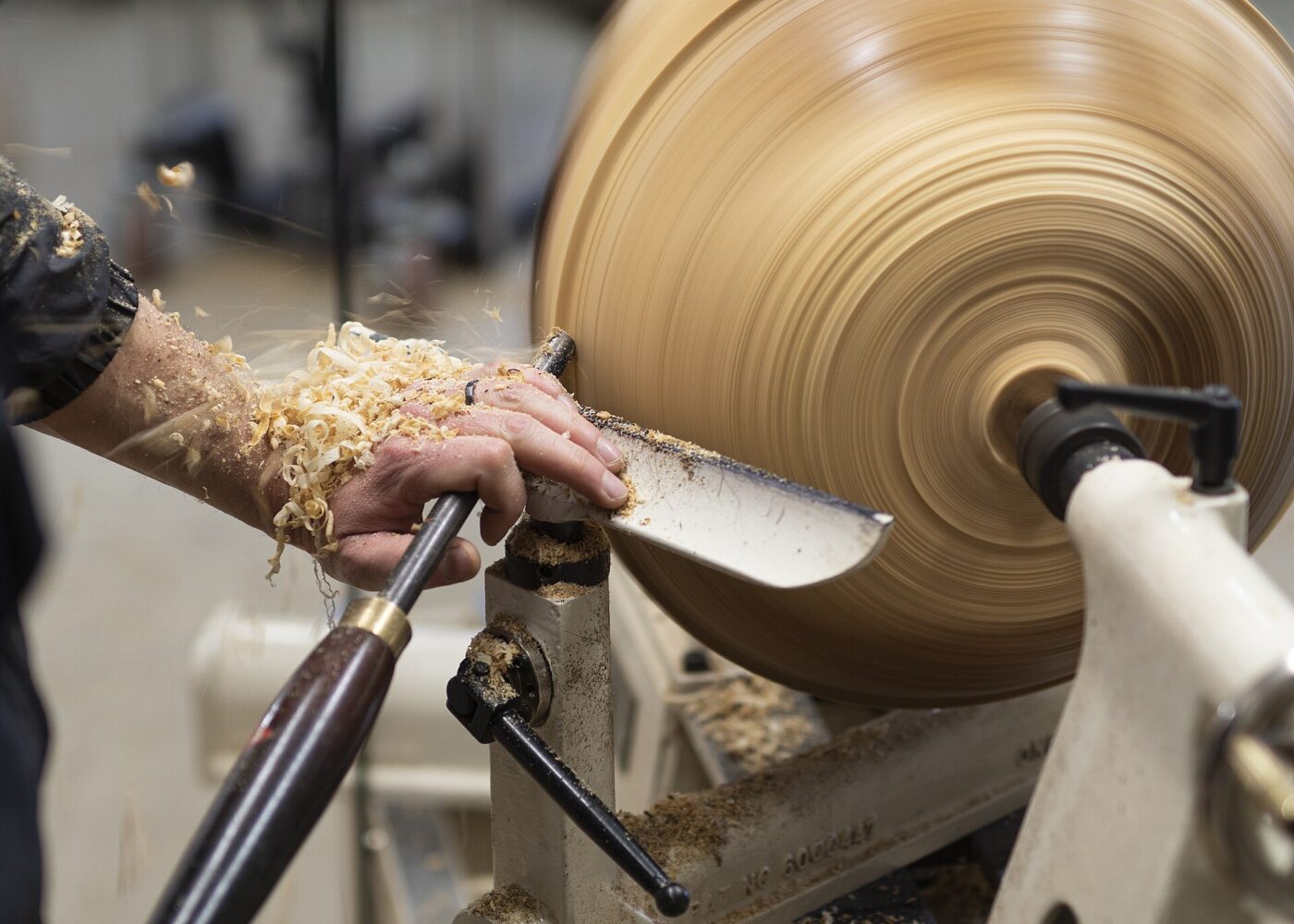A wood bowl blank spins in a blur on a lathe while a hand guides a gouge through flying shavings.