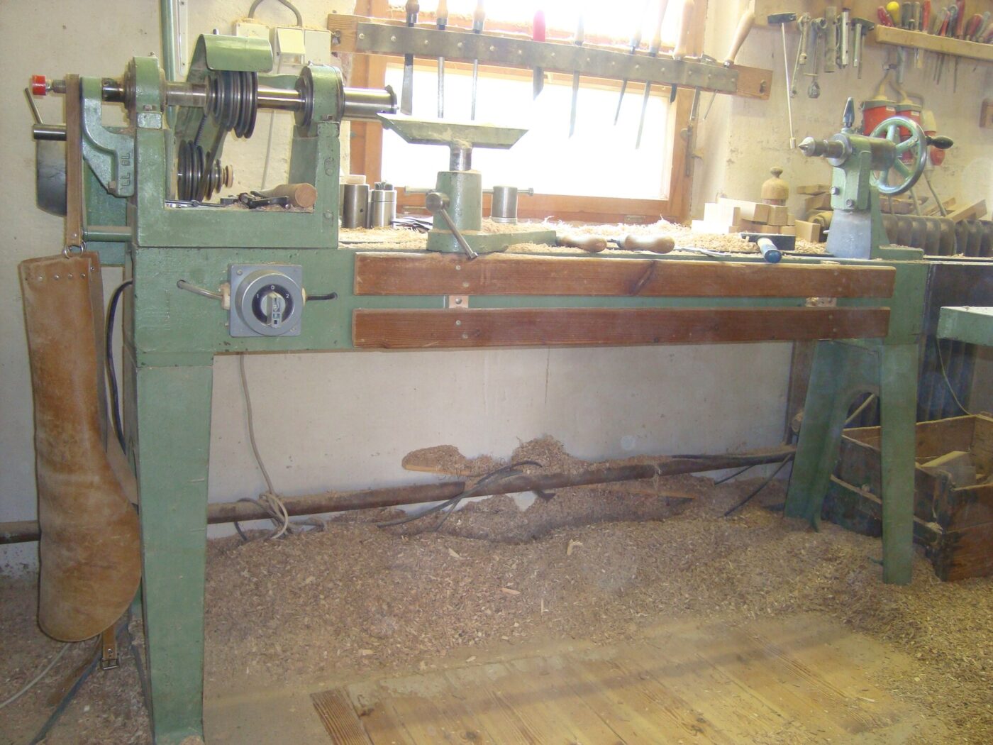 A green wood lathe with a long bed stands in a wood shop, surrounded by shavings.