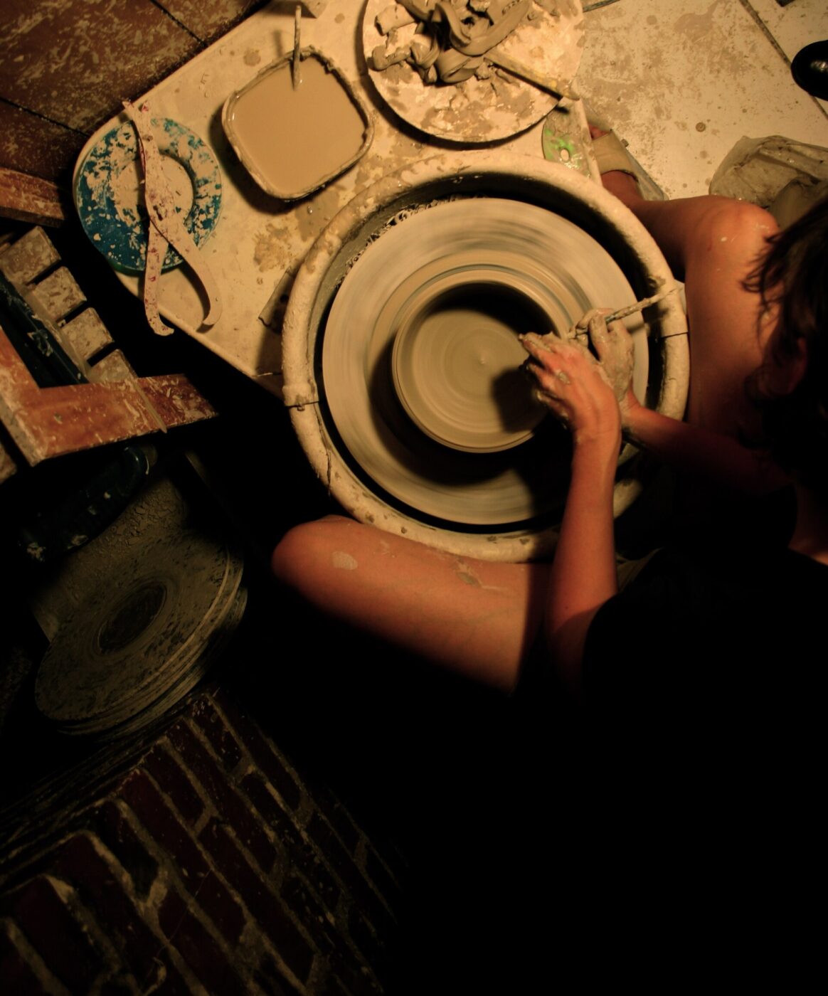 An overhead view of a potter's hands shaping a bowl on a spinning electric pottery wheel surrounded by tools.