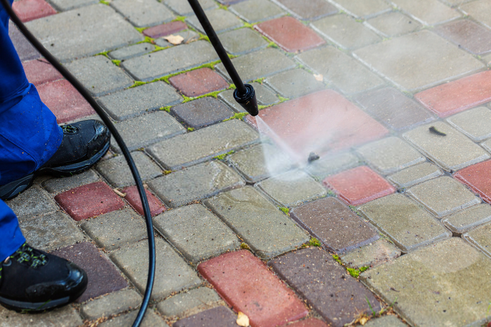 A pressure washer wand cleaning a brick paver driveway.