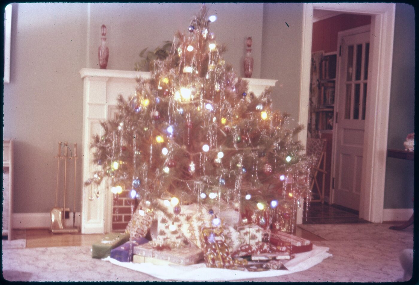 A decorated, lit Christmas tree and wrapped presents beside a white fireplace mantel indoors.