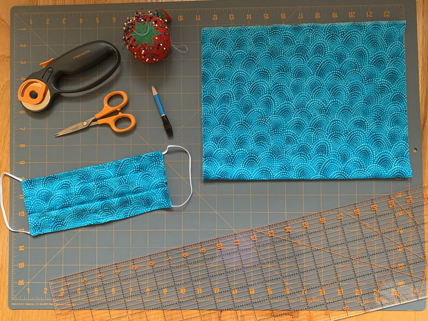 An orange rotary cutter, scissors, a pincushion and thread rest on a gridded cutting mat beside blue fabric.