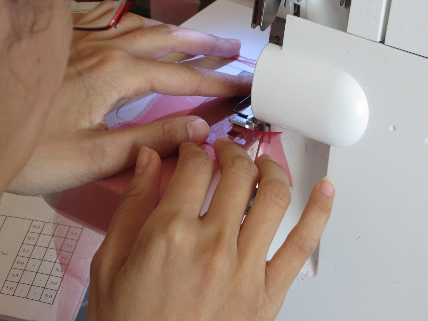 A close view of hands feeding pink fabric into a white serger sewing machine.
