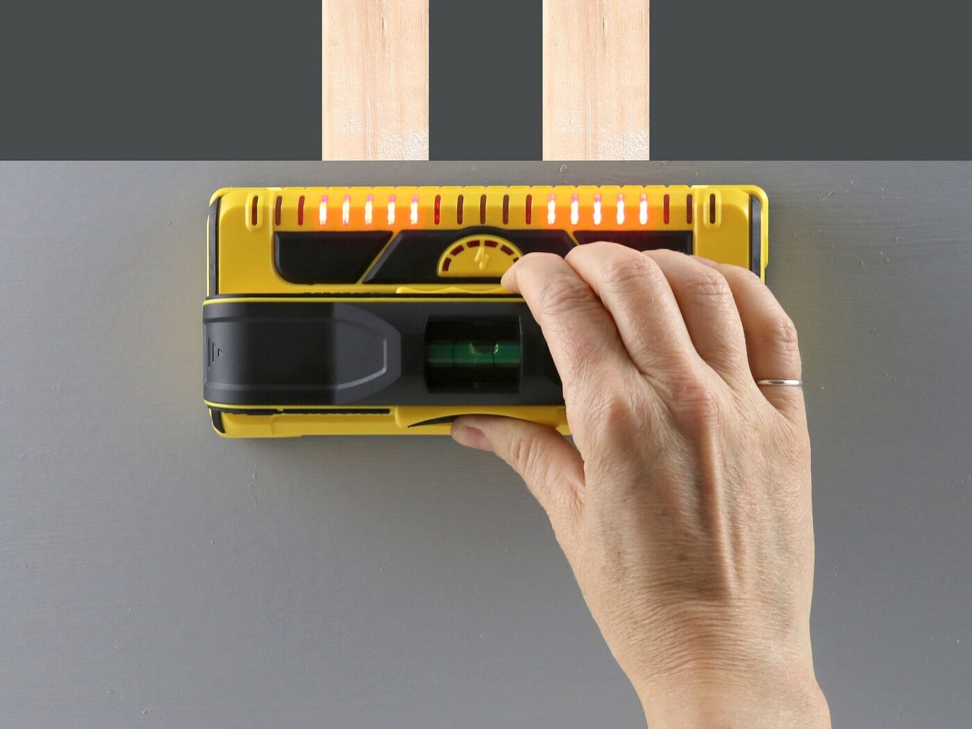 A hand holds an electronic stud finder against a wall, its indicator lights lit.