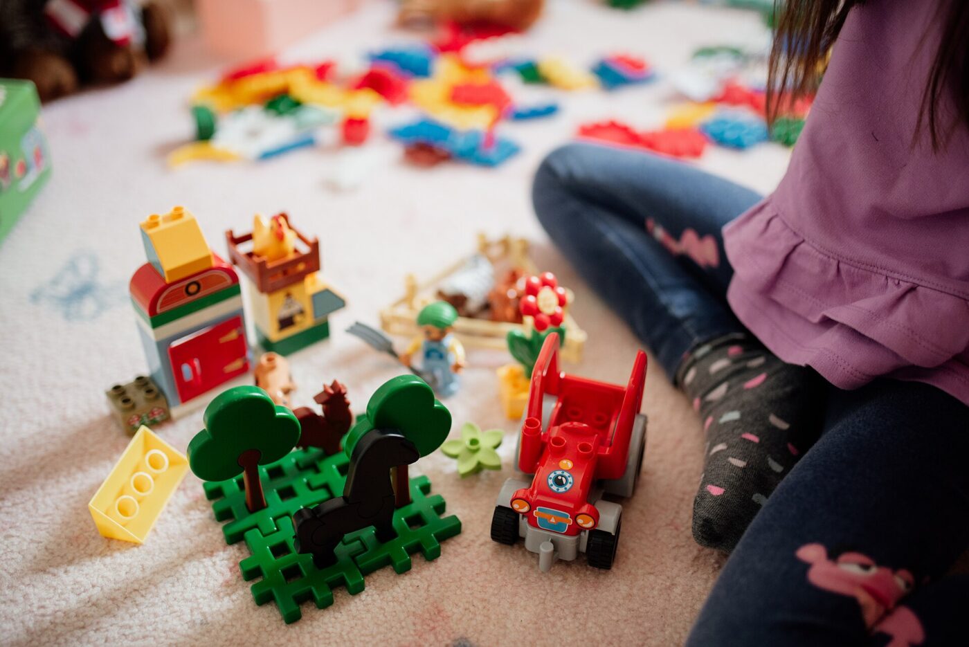 Small colorful toy blocks and figures scattered on a floor near a young child's legs and feet.