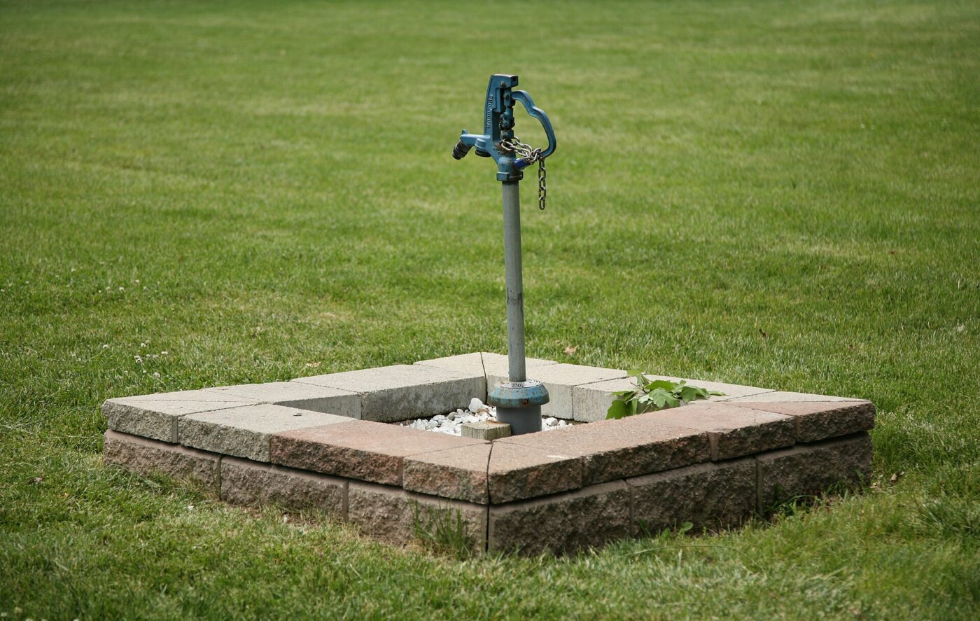 Metal hand-pump wellhead mounted on a square concrete surround in grass.